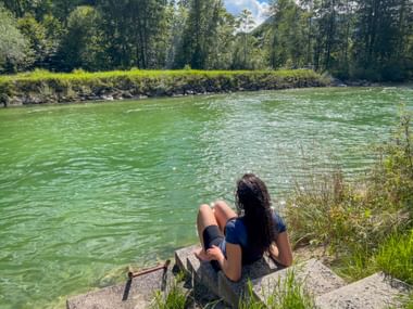 Woman with long dark hair sitting on a rock by the turquoise Ischler Ache river in Salzkammergut, surrounded by green meadows and trees.