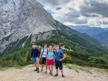 Four hikers with backpacks posing at Vršič Pass in the Julian Alps. Behind them, dramatic mountain peaks and forested slopes under cloudy skies.