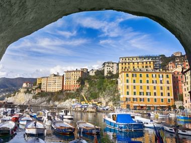 View of Camogli harbor through a stone arch, showing colorful buildings on a hillside, fishing boats in the water, and blue sky above.