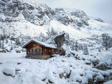 Holzhütte mit Steinfundament im verschneiten Obersulzbachtal, Hohe Tauern. Schneebedeckte Felsen und Berge umgeben die Hütte.