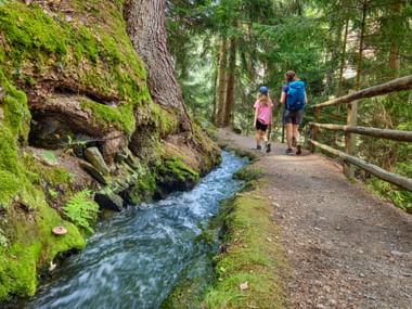 Zwei Wanderer auf einem Waldweg neben einem fließenden Bach in Schluderns, Südtirol. Moosbewachsene Felsen und Holzgeländer säumen den Weg.