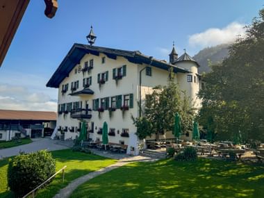 White castle building with green shutters and tower in Pinzgau. Garden terrace with tables and umbrellas on manicured lawn, mountains in background.
