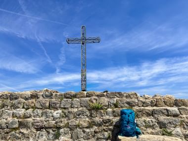 Metallenes Gipfelkreuz auf felsigem Gipfel La Rocca mit blauem Rucksack am Fuß. Klarer blauer Himmel mit Schleierwolken über der Gardasee-Region.