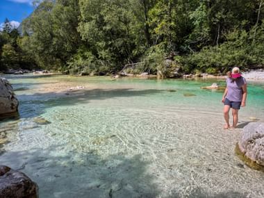 Female hiker with pink cap standing on rock in crystal-clear turquoise Soča River in Julian Alps, surrounded by forest and rocky riverbed.
