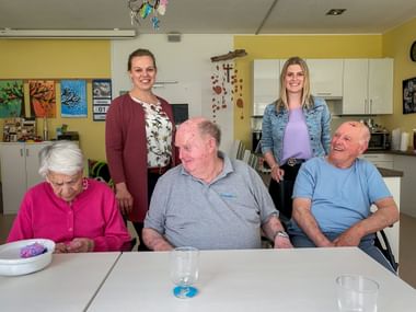 Zwei Mitarbeiterinnen stehen hinter drei sitzenden Senioren an einem Tisch in einem hellen Gemeinschaftsraum mit gelben Wänden und bunten Bildern.