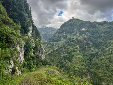 Narrow hiking trail along steep cliffs on Madeira's north coast, with lush green mountains and white buildings visible in the distance.