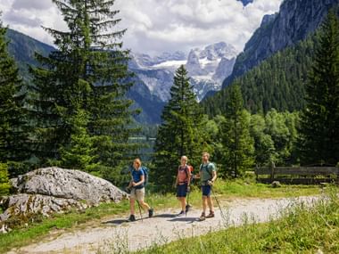 Three hikers with backpacks walking on a mountain path near Gosausee with snow-capped peaks, green forests, and a lake in the background.