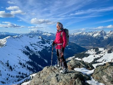 Wanderer in pinker Jacke mit Rucksack und Stöcken auf felsigem Gipfel der Schwalbenwand, umgeben von schneebedeckten Alpengipfeln unter blauem Himmel.
