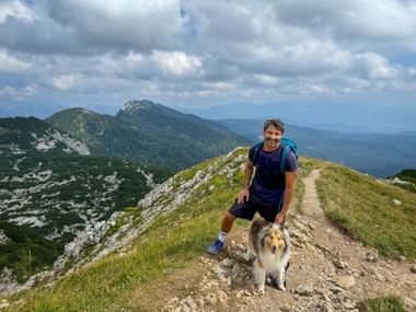 Smiling hiker with backpack kneeling beside a collie dog on a mountain trail in the Julian Alps with green peaks and cloudy sky.