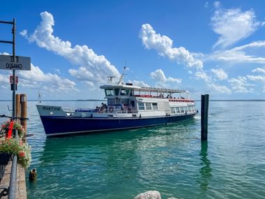 Blau-weißes Fährboot namens Mantova im Hafen von Dusano am Gardasee. Türkisfarbenes Wasser und blauer Himmel mit weißen Wolken sichtbar.