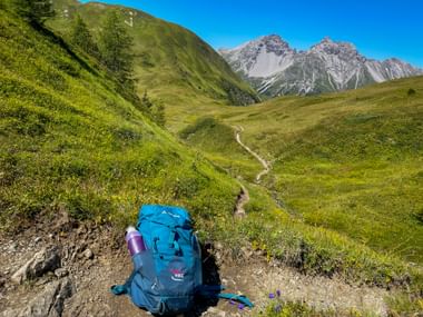 Blauer Wanderrucksack auf einem Bergweg mit grünen Almwiesen und felsigen Gipfeln der Allgäuer Alpen unter klarem Himmel.