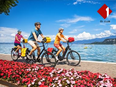 Drei Radfahrer mit bunten Packtaschen fahren entlang einer Seepromenade mit Blumen, Bergen und Ö1 Club Logo.