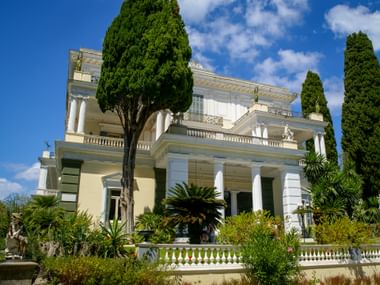 Achilleion Palace, a neoclassical building with white columns and balustrades, surrounded by cypress trees and Mediterranean vegetation under blue sky.