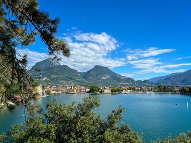 Blick auf Riva del Garda am Gardasee mit Bergen im Hintergrund. Kiefernzweige rahmen die Szene mit türkisfarbenem Wasser und blauem Himmel.