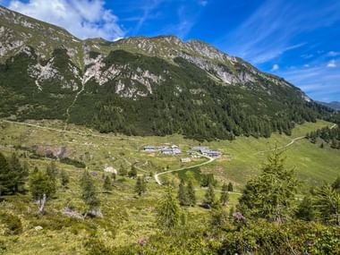 Bergsiedlung mit traditionellen Hütten auf grünen Almwiesen unterhalb bewaldeter Gipfel. Gewundene Wege verbinden die Gebäude im Tal.