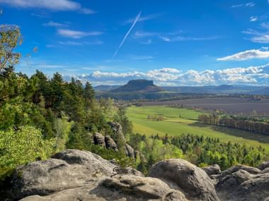 View from Rauenstein rock formations across green fields and forests to the distinctive table mountain Lilienstein under blue sky with clouds.