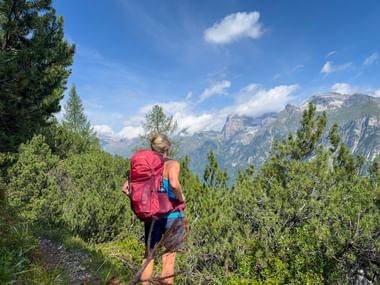 Wanderin mit rotem Rucksack auf Bergpfad mit Blick auf Alpengipfel bei Ladurns, umgeben von grünen Kiefern unter blauem Himmel.