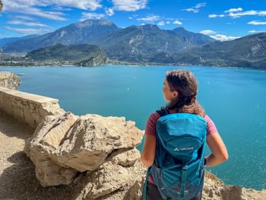 Wanderin mit blauem Rucksack blickt vom Felsklippe auf türkisfarbenen Gardasee, mit Bergen und blauem Himmel im Hintergrund.