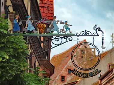 Kunstvolles schmiedeeisernes Zunftschild der Weinkellerei Hugel im Elsass mit bemalten Figuren in Tracht und rundem Emblem mit Traubenmotiven.