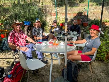 Gruppe von fünf Wanderern sitzt um einen runden Tisch in einem sonnigen toskanischen Garten bei Montecchio und genießt eine Pause mit Kaffee.