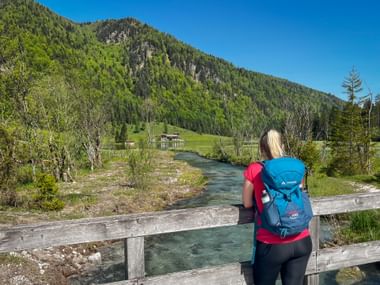 Wanderin mit blauem Rucksack auf Holzbrücke mit Blick auf türkisfarbenen Bach am Pillersee, mit bewaldeten Bergen in den Kitzbüheler Alpen.