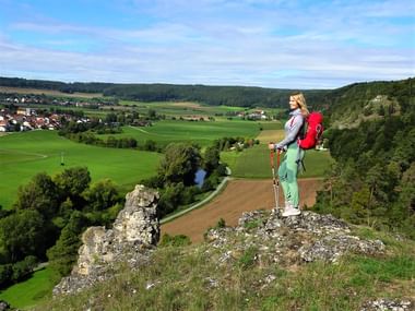 Wanderin mit rotem Rucksack steht auf Felsvorsprung mit Blick über das Altmühltal mit grünen Feldern, Dorf und bewaldeten Hügeln.