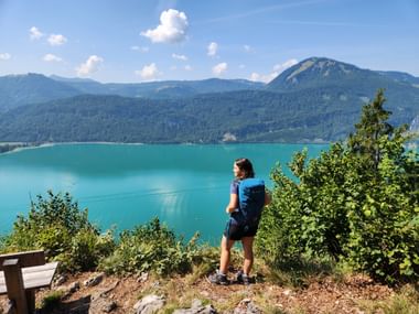 Wanderin mit Rucksack blickt auf den türkisfarbenen Wolfgangsee im Salzkammergut, Österreich. Berge und Wälder umgeben den See unter blauem Himmel.