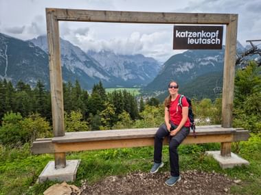 Wanderin in rotem Shirt sitzt auf Holzbank des Panoramarahmens am Katzenkopf Leutasch mit Blick auf Bergtal und Zugspitzregion.
