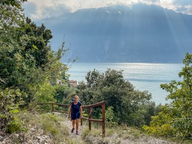 Wanderin auf dem Sentiero del Sole mit Gardasee und Bergen im Hintergrund, umgeben von mediterraner Vegetation und Holzzaun.