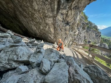 Woman crouching on rocky path under massive limestone overhang in Rosengartenschlucht near Imst, with wooden fence and valley view.