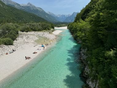 Türkisfarbener Isonzo mit Kiesstrand bei Bovec. Menschen entspannen am Ufer, umgeben von grünen Wäldern und Alpenbergen.