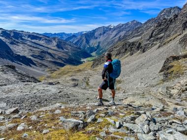 Wanderer mit blauem Rucksack auf felsigem Bergpfad mit Blick auf das Pfossental. Alpine Gipfel und grünes Tal in der Ferne unter blauem Himmel.