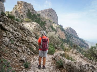 Hiker with red backpack on rocky mountain trail facing dramatic limestone peaks of Cornador Gran on the Trans Tramuntana route.