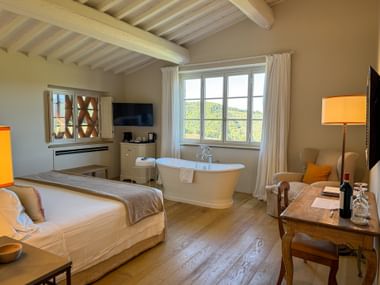 Geräumiges Zimmer im Hotel Pietra del Cabreo mit Balkendecke, freistehender Badewanne am Fenster, Doppelbett und Blick auf toskanische Weinberge.