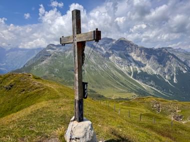 Hölzernes Gipfelkreuz am Sandjöchl mit Panoramablick auf Alpengipfel und grüne Täler unter blauem Himmel mit weißen Wolken.