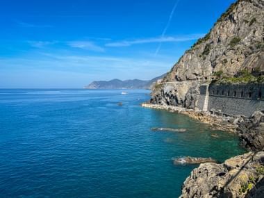 Dramatic coastal path carved into rocky cliffs in Cinque Terre, Liguria. Turquoise Mediterranean Sea and distant mountains under blue sky.