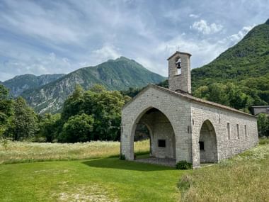 Stone chapel with bell tower and arched openings in Venzone, Friuli, set against green mountains and cloudy sky in a grassy meadow.