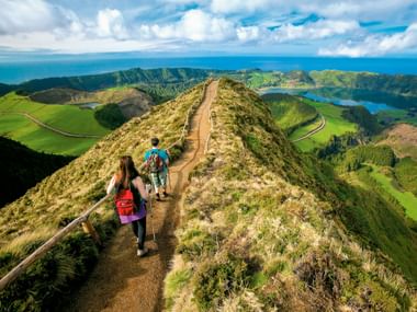 Zwei Wanderer auf schmalem Grat am Kraterrand von Sete Cidades auf den Azoren, mit Vulkanseen und grünen Hügeln unter blauem Himmel.