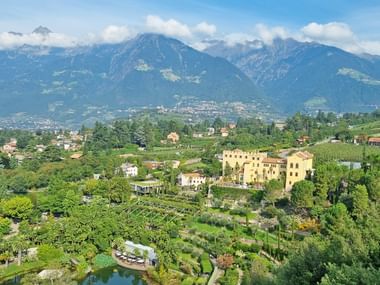 Aerial view of Trauttmansdorff Castle with terraced botanical gardens in South Tyrol, surrounded by mountains and valley settlements.