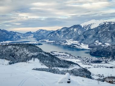 Luftaufnahme des Wolfgangsees im Winter, umgeben von schneebedeckten Bergen und Wäldern. Dörfer säumen das Ufer unter bewölktem Himmel.