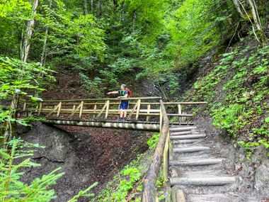 Wanderer überquert eine Holzbrücke auf einem Serpentinenweg im Wald bei Garmisch, umgeben von üppiger grüner Vegetation und steilen Hängen.