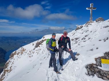 Zwei Wanderer in Winterausrüstung auf dem verschneiten Schafberg Gipfel mit Metallkreuz und Bergpanorama unter blauem Himmel.