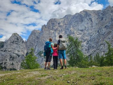 Drei Wanderer mit Rucksäcken betrachten dramatische Felsgipfel in den Julischen Alpen beim Vršič-Pass. Grüne Wiese und Kiefern im Vordergrund.