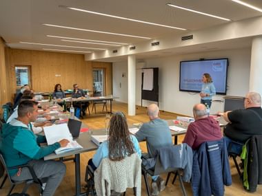 Training session in modern conference room with presenter at screen showing 'Willkommen' and participants seated at tables with materials.