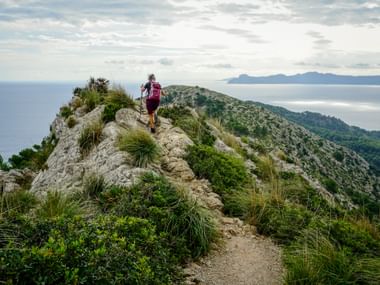 Hiker with pink backpack on rocky mountain trail near Alcudia, overlooking Mediterranean Sea and distant mountains under cloudy sky.
