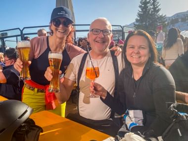 Drei Personen genießen Getränke auf einer sonnigen Bergterrasse beim Wintersporttag 2026, mit schneebedeckten Gipfeln und Tannen im Hintergrund.