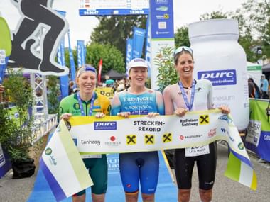 Three female triathletes holding a course record banner at Trumer Triathlon finish line, wearing medals and race gear, smiling at camera.
