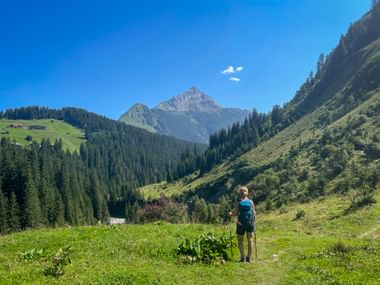 Wanderin mit Rucksack und Wanderstöcken steht auf grüner Almwiese mit Blick auf Berggipfel in den Allgäuer Alpen unter blauem Himmel.