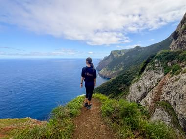 Female hiker walking on Vereda do Larano coastal trail in Madeira with steep cliffs, green vegetation, and deep blue Atlantic Ocean.