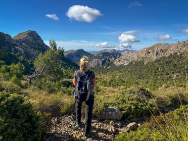 Hiker with backpack standing on rocky trail viewing Tramuntana mountain range under blue sky with scattered clouds and green vegetation.